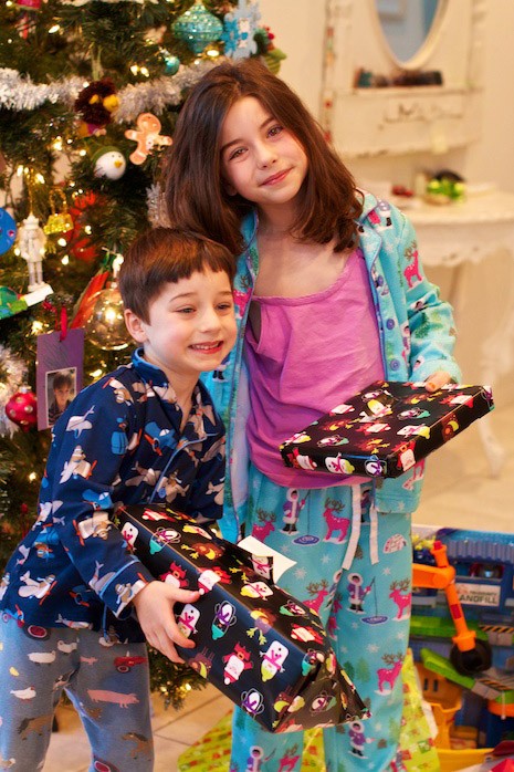 Little kids in pajamas opening Christmas presents in front of holiday tree.
